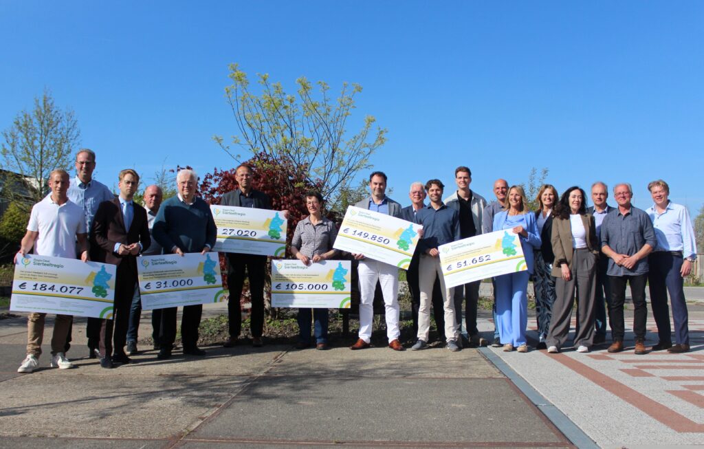 Foto van alle betrokkenen op een rijtje, buiten, inclusief de 6 vertegenwoordigers met een grote cheque in hun handen, met daarop het gegunde bedrag voor hun project.